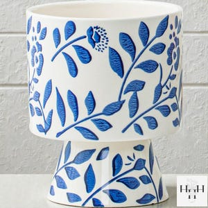 Puede incluir: Jardinera de cer&aacute;mica blanca con base de pedestal, adornada con un patr&oacute;n floral azul. El dise&ntilde;o presenta hojas y flores estilizadas, creando un aspecto decorativo y elegante. Perfecto para plantas de interior.