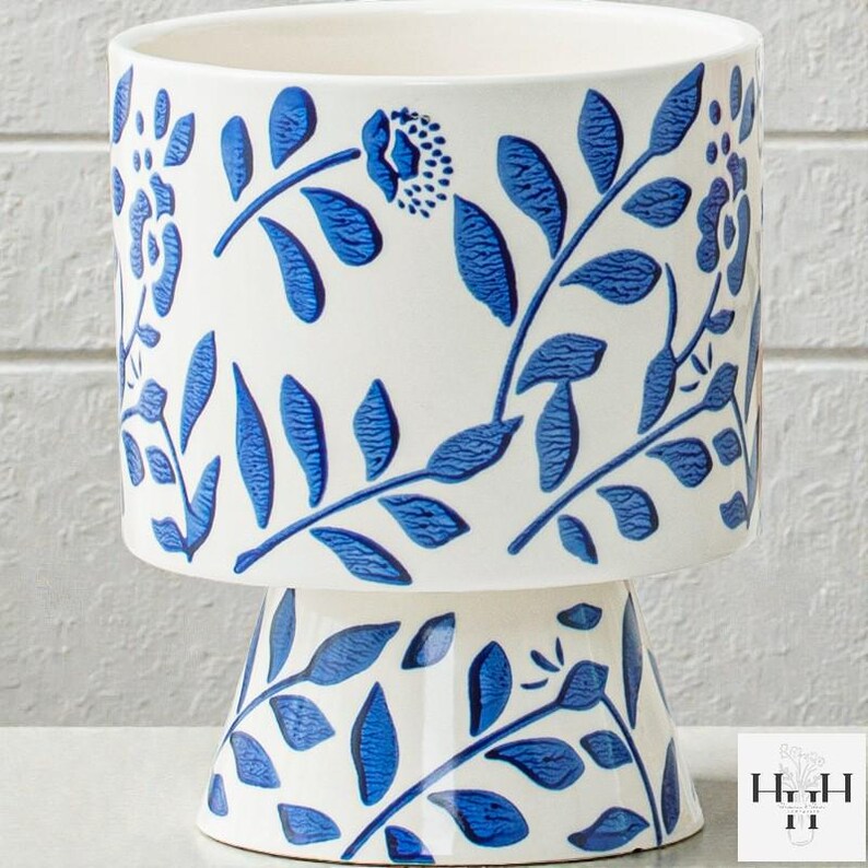 Puede incluir: Jardinera de cer&aacute;mica blanca con base de pedestal, adornada con un patr&oacute;n floral azul. El dise&ntilde;o presenta hojas y flores estilizadas, creando un aspecto decorativo y elegante. Perfecto para plantas de interior.