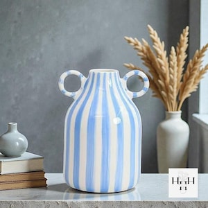May include: A ceramic vase with a blue and white striped pattern. The vase has two loop handles and a wide opening. A small gray vase and a stack of books are to the left. Dried wheat stalks are in a white vase to the right.