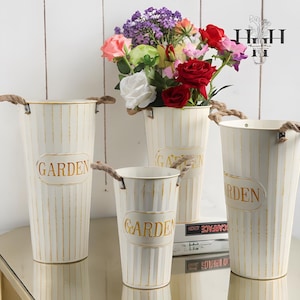 Könnte beinhalten: Vier cremefarbene Gartenvasen aus Metall mit Seilgriffen. Die Vasen haben vertikale Streifen und das Wort "GARDEN" in goldfarbenen Buchstaben. Eine Vase enthält einen Strauß bunter Kunstblumen.