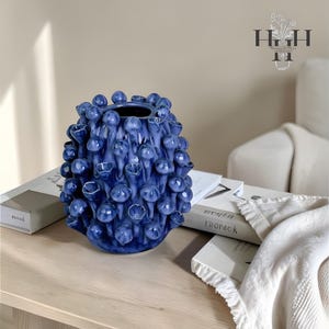 May include: A cobalt blue ceramic vase with a textured, organic design. The vase features a series of rounded, bud-like shapes and open, flower-like forms. The vase is displayed on a light-colored wooden surface, with books and a white throw in the background.