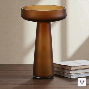 May include: A brown glass vase with a wide, shallow top and a conical base. The vase has a frosted finish and sits on a wooden surface. The overall design is modern and minimalist, suitable for home decor.
