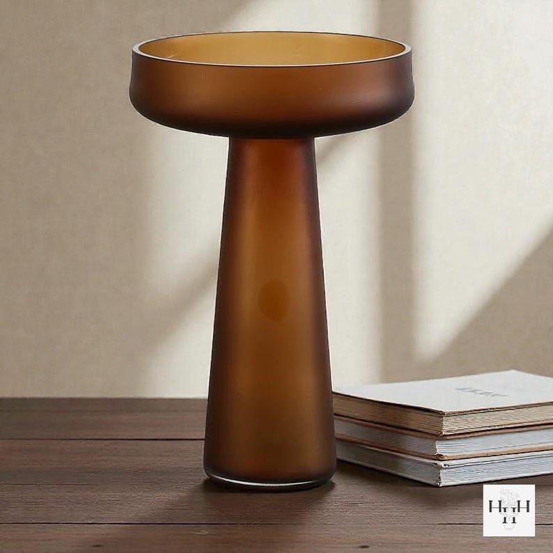 May include: A brown glass vase with a wide, shallow top and a conical base. The vase has a frosted finish and sits on a wooden surface. The overall design is modern and minimalist, suitable for home decor.