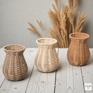 Puede incluir: Tres jarrones de mimbre tejido en tonos marrones y beige sobre una superficie de madera clara. Los jarrones tienen forma redondeada con la parte superior ensanchada. Tallos de trigo seco en el fondo.