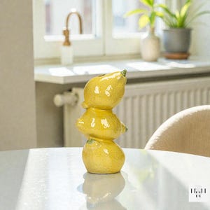 Puede incluir: Un jarrón de cerámica con forma de tres limones amarillos apilados. El jarrón tiene un acabado brillante y está sobre una mesa blanca. Los limones tienen tallos verdes y están dispuestos verticalmente.