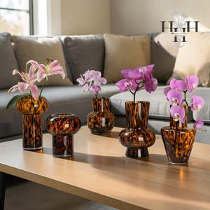 May include: Five decorative glass vases with a tortoiseshell pattern, each holding fresh flowers. The vases vary in shape and size, with a warm brown and amber colour scheme. The flowers include pink lilies and purple orchids, arranged on a wooden table.