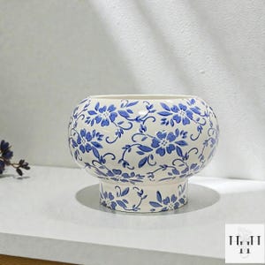 Puede incluir: Una maceta de cer&aacute;mica blanca con un estampado floral azul. La maceta redonda se asienta sobre una base de pedestal, y el dise&ntilde;o floral azul cubre toda la superficie. La maceta es adecuada para uso en interiores o exteriores.