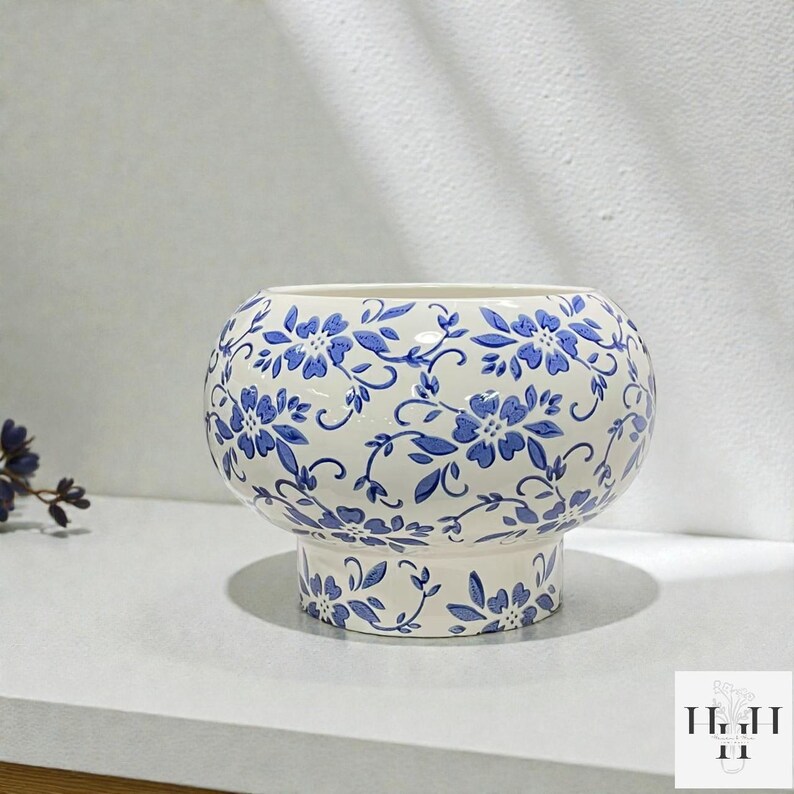 Puede incluir: Una maceta de cer&aacute;mica blanca con un estampado floral azul. La maceta redonda se asienta sobre una base de pedestal, y el dise&ntilde;o floral azul cubre toda la superficie. La maceta es adecuada para uso en interiores o exteriores.