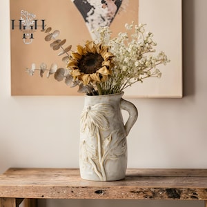 May include: A cream-colored ceramic pitcher with a handle, adorned with embossed floral designs. The pitcher holds a bouquet of dried flowers, including a sunflower and delicate white blooms. The pitcher is approximately 10 inches tall.