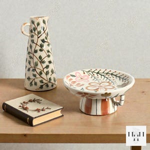 May include: A ceramic jug with a handle, decorated with green leaves and brown branches on a cream background. A small, round, pedestal dish with a floral design and a book with a floral design are also present.