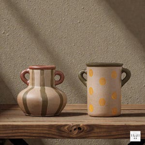 Puede incluir: Dos jarrones de cerámica con asas sobre una estantería de madera. Uno es beige con rayas marrones y un borde marrón. El otro es beige con lunares amarillos y un borde verde. Ambos tienen un aspecto rústico y hecho a mano.