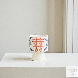Puede incluir: Una maceta de cer&aacute;mica blanca con base de pedestal. El cuerpo cil&iacute;ndrico presenta un dise&ntilde;o rojo y azul con un s&iacute;mbolo de doble felicidad y detalles florales. La maceta es adecuada para plantas peque&ntilde;as o uso decorativo.