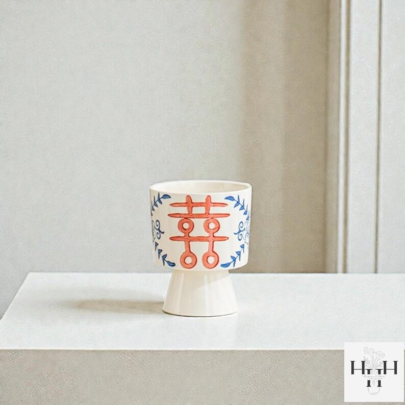 Puede incluir: Una maceta de cer&aacute;mica blanca con base de pedestal. El cuerpo cil&iacute;ndrico presenta un dise&ntilde;o rojo y azul con un s&iacute;mbolo de doble felicidad y detalles florales. La maceta es adecuada para plantas peque&ntilde;as o uso decorativo.