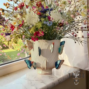 Könnte beinhalten: Eine cremefarbene Vase mit Schmetterlingsakzenten hält einen lebendigen Strauß bunter Blumen. Die Vase hat eine blattartige Textur und Ausschnitte. Die Schmetterlinge sind in Blau-, Rosa- und Rottönen gehalten.
