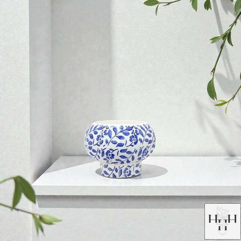 Puede incluir: Un jarr&oacute;n de cer&aacute;mica blanco y azul con un dise&ntilde;o floral. El jarr&oacute;n tiene un cuerpo redondeado y una base de pedestal. El dise&ntilde;o floral azul cubre toda la superficie. El jarr&oacute;n est&aacute; sobre una estanter&iacute;a blanca.