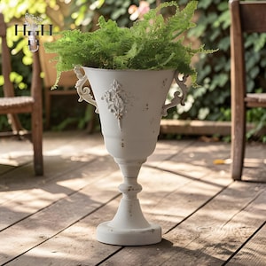 May include: A cream-coloured, distressed urn-shaped planter with decorative handles and a front panel. The planter contains a vibrant green fern. The planter sits on a wooden deck, with a blurred background of greenery and wooden chairs.
