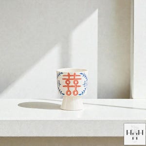 Puede incluir: Una maceta de cer&aacute;mica blanca con base de pedestal. La maceta presenta un dise&ntilde;o rojo y azul con un s&iacute;mbolo de doble felicidad y detalles florales. El dise&ntilde;o est&aacute; pintado a mano en el exterior de la maceta.
