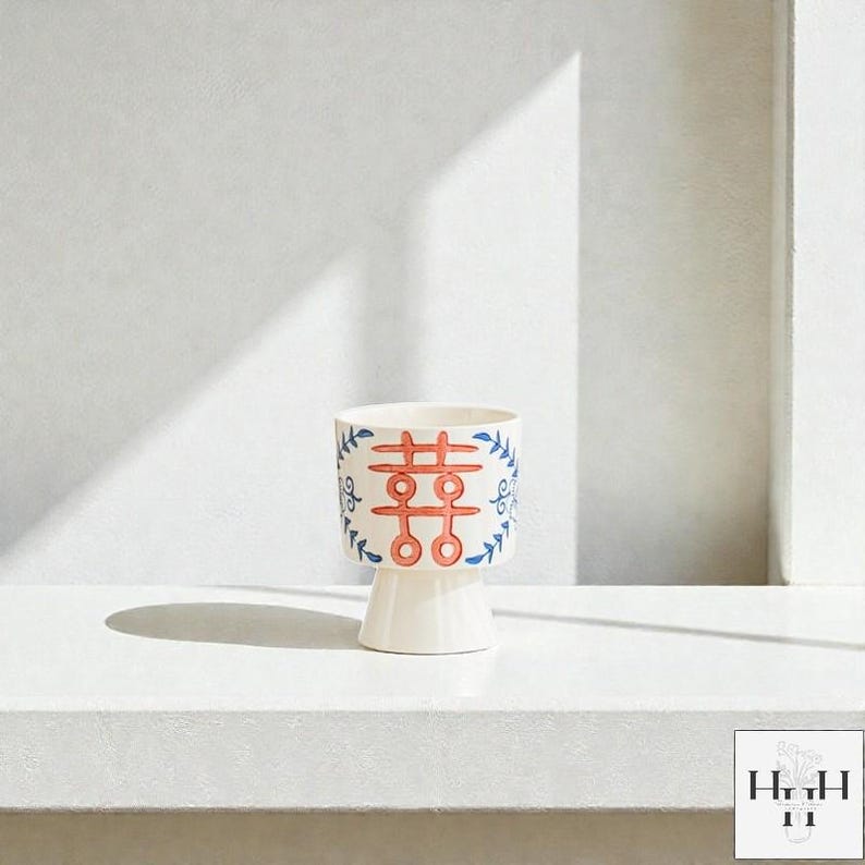 Puede incluir: Una maceta de cer&aacute;mica blanca con base de pedestal. La maceta presenta un dise&ntilde;o rojo y azul con un s&iacute;mbolo de doble felicidad y detalles florales. El dise&ntilde;o est&aacute; pintado a mano en el exterior de la maceta.