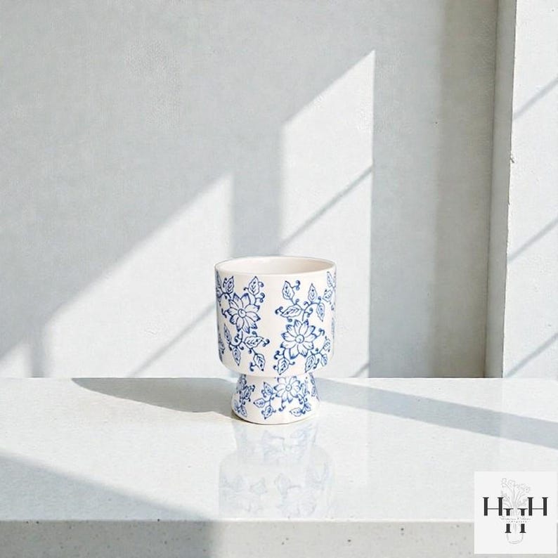 Puede incluir: Una maceta de cer&aacute;mica blanca con un estampado floral azul. La maceta tiene una base de pedestal y una parte superior cil&iacute;ndrica. El dise&ntilde;o floral incluye flores y hojas. La maceta est&aacute; sobre una superficie blanca.