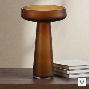May include: A brown glass vase with a wide, shallow top and a conical base. The vase has a frosted finish and sits on a dark wooden surface. A stack of books is next to the vase. The vase is a decorative home decor item.
