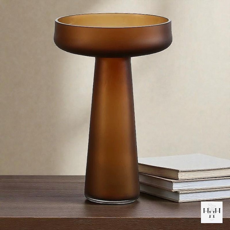 May include: A brown glass vase with a wide, shallow top and a conical base. The vase has a frosted finish and sits on a dark wooden surface. A stack of books is next to the vase. The vase is a decorative home decor item.