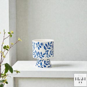 Puede incluir: Una maceta de cer&aacute;mica blanca con un dise&ntilde;o floral azul. La maceta tiene una parte superior cil&iacute;ndrica y una base de pedestal, ambas adornadas con hojas y flores azules. El dise&ntilde;o se repite alrededor de toda la maceta.