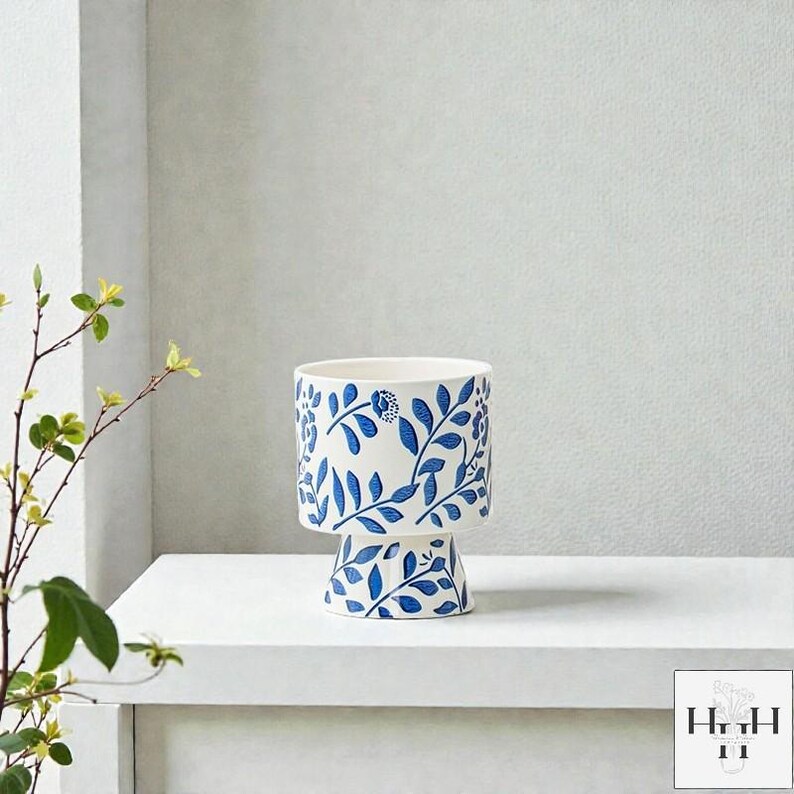 Puede incluir: Una maceta de cer&aacute;mica blanca con un dise&ntilde;o floral azul. La maceta tiene una parte superior cil&iacute;ndrica y una base de pedestal, ambas adornadas con hojas y flores azules. El dise&ntilde;o se repite alrededor de toda la maceta.
