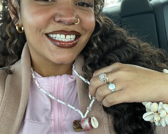 May include: A woman wearing a pink zip-up jacket and a white beaded necklace with a small glass vial pendant. The pendant is decorated with pink and white accents and a seashell. She is also wearing rings and a bracelet with seashells.