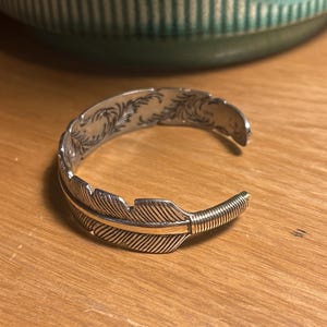 May include: Silver cuff bracelet with a feather design. The bracelet has detailed feather etching and a gold-coloured accent. The open cuff design allows for easy wear. The bracelet is displayed on a wooden surface.