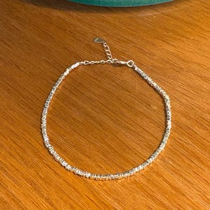 Puede incluir: Una delicada pulsera de plata con un pequeño dije de corazón. La pulsera está hecha de pequeñas cuentas espaciadas uniformemente y se muestra sobre una superficie de madera. La pulsera es ajustable.