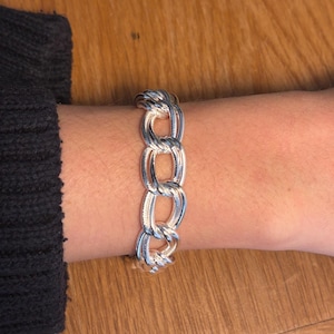 May include: A silver-colored chain link bracelet. The bracelet is made of interlocking oval links. The bracelet is worn on a wrist, with a dark gray sleeve visible in the background. The bracelet is a classic jewelry piece.