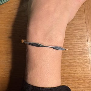 Puede incluir: Una pulsera de puño plateada con un diseño retorcido. La pulsera tiene un acabado liso y pulido y se lleva en la muñeca. El diseño de la pulsera presenta un sutil patrón ondulado, que añade un toque de elegancia a la joya.