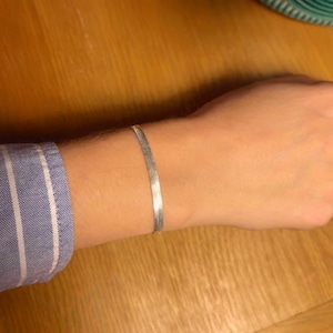 May include: A silver herringbone bracelet is displayed on a wrist. The bracelet is composed of small, flat, interlocking metal pieces. The person is wearing a blue and white striped shirt. The background is a wooden surface.