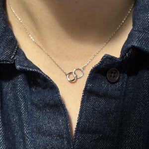 Könnte beinhalten: Eine silberne Halskette mit zwei ineinandergreifenden Kreisen, einer davon mit kleinen, funkelnden Kristallen besetzt. Die feine Kette und der Anhänger heben sich vor einem dunkelblauen Jeanshemd ab und betonen die Eleganz des Schmucks.