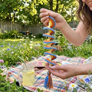 May include: A colorful, spiral-shaped crochet hanging decoration. The handmade piece features a repeating pattern of orange, yellow, and blue. A blue and orange tassel hangs from the bottom. The background shows a picnic setting with a drink.