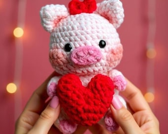 Könnte beinhalten: Ein handgehäkeltes Ferkel in Rosa- und Weißtönen, das ein rotes Herz hält. Das Ferkel hat eine kleine rote Schleife auf dem Kopf und schwarze Knopfaugen. Das Stofftier wird vor einem rosa Hintergrund gehalten.