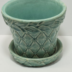 May include: A teal ceramic planter with a matching saucer. The planter has a quilted pattern and a decorative leaf design around the rim. The planter and saucer are made of ceramic.