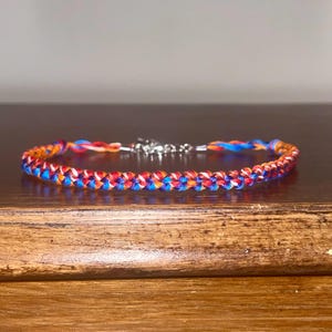 May include: A braided bracelet featuring a vibrant mix of red, blue, and orange threads. The bracelet has a silver-colored clasp. The braided design is visible, showcasing the interwoven strands of color. This accessory is suitable for everyday wear.