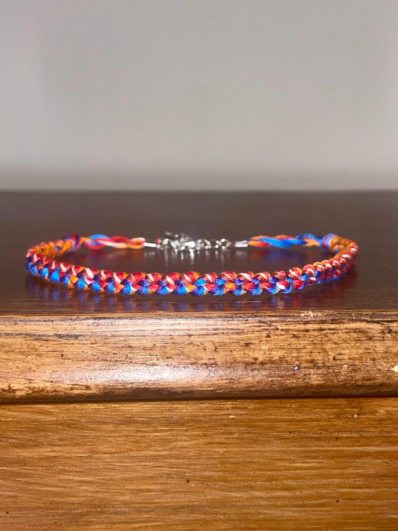 May include: A braided bracelet featuring a vibrant mix of red, blue, and orange threads. The bracelet has a silver-colored clasp. The braided design is visible, showcasing the interwoven strands of color. This accessory is suitable for everyday wear.