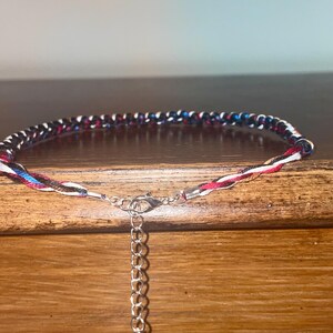 May include: A braided anklet with a mix of red, white, blue, and black threads. The anklet has a silver-colored clasp and an adjustable chain. The design is simple and colorful, suitable for casual wear.