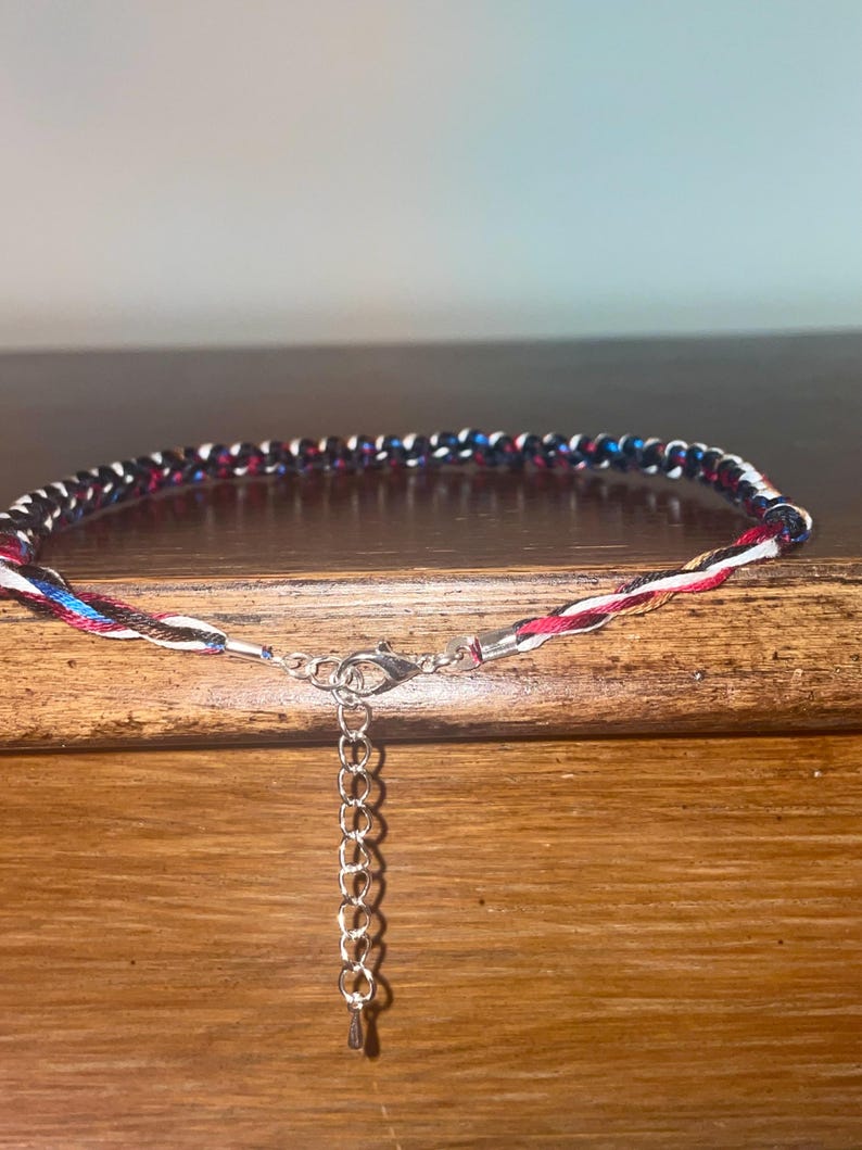 May include: A braided anklet with a mix of red, white, blue, and black threads. The anklet has a silver-colored clasp and an adjustable chain. The design is simple and colorful, suitable for casual wear.