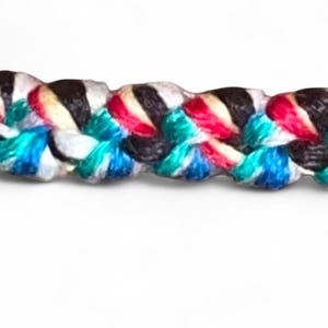May include: Close-up of a braided rope with a vibrant mix of colors. The rope features strands of black, white, red, turquoise, and blue, creating a textured and visually appealing pattern. The background is plain white.