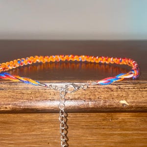 May include: Braided bracelet featuring orange, red, blue, and yellow threads. The bracelet has a silver-colored clasp and an adjustable chain. The bracelet is displayed on a wooden surface.