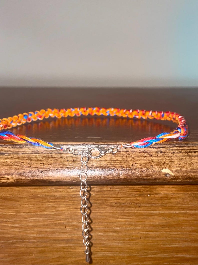 May include: Braided bracelet featuring orange, red, blue, and yellow threads. The bracelet has a silver-colored clasp and an adjustable chain. The bracelet is displayed on a wooden surface.