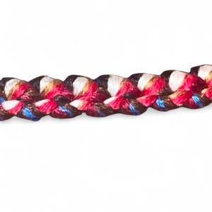 May include: Close-up of a braided cord with a repeating pattern of red, white, blue, and black threads. The cord is tightly woven, creating a textured surface. The colors are vibrant against a white background.