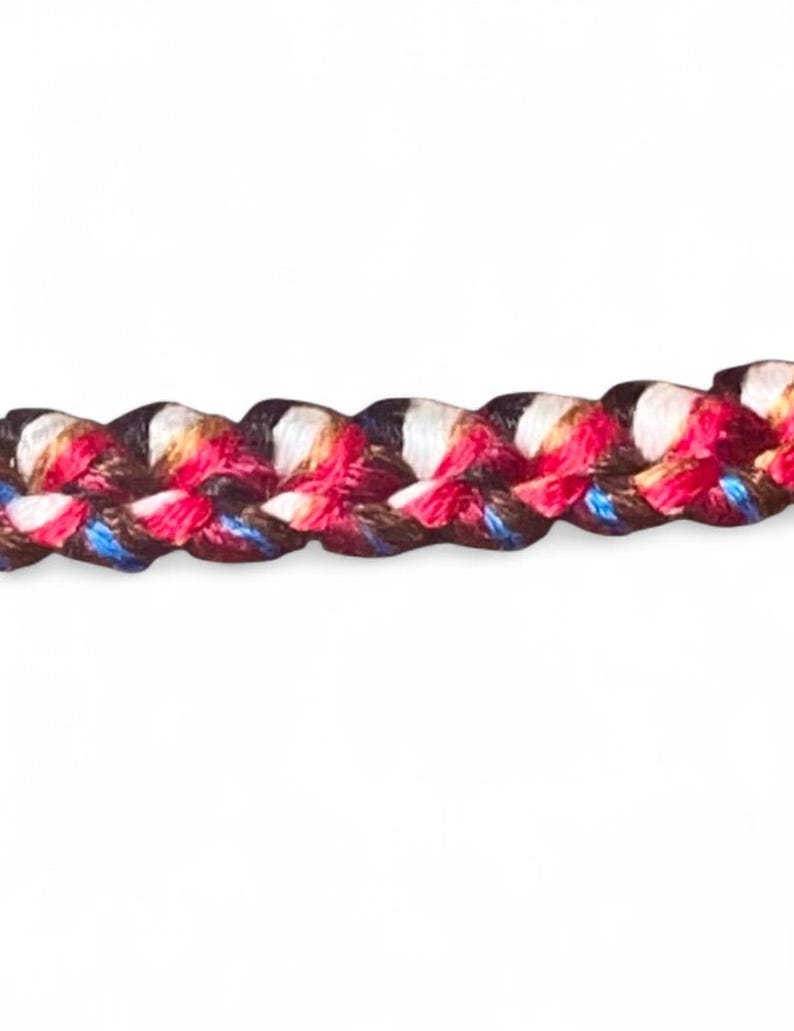 May include: Close-up of a braided cord with a repeating pattern of red, white, blue, and black threads. The cord is tightly woven, creating a textured surface. The colors are vibrant against a white background.