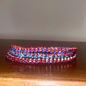 May include: A stack of colorful braided bracelets. The bracelets are made of red, white, blue, and orange threads. The bracelets are stacked on top of each other, creating a layered effect. The bracelets are on a wooden surface.