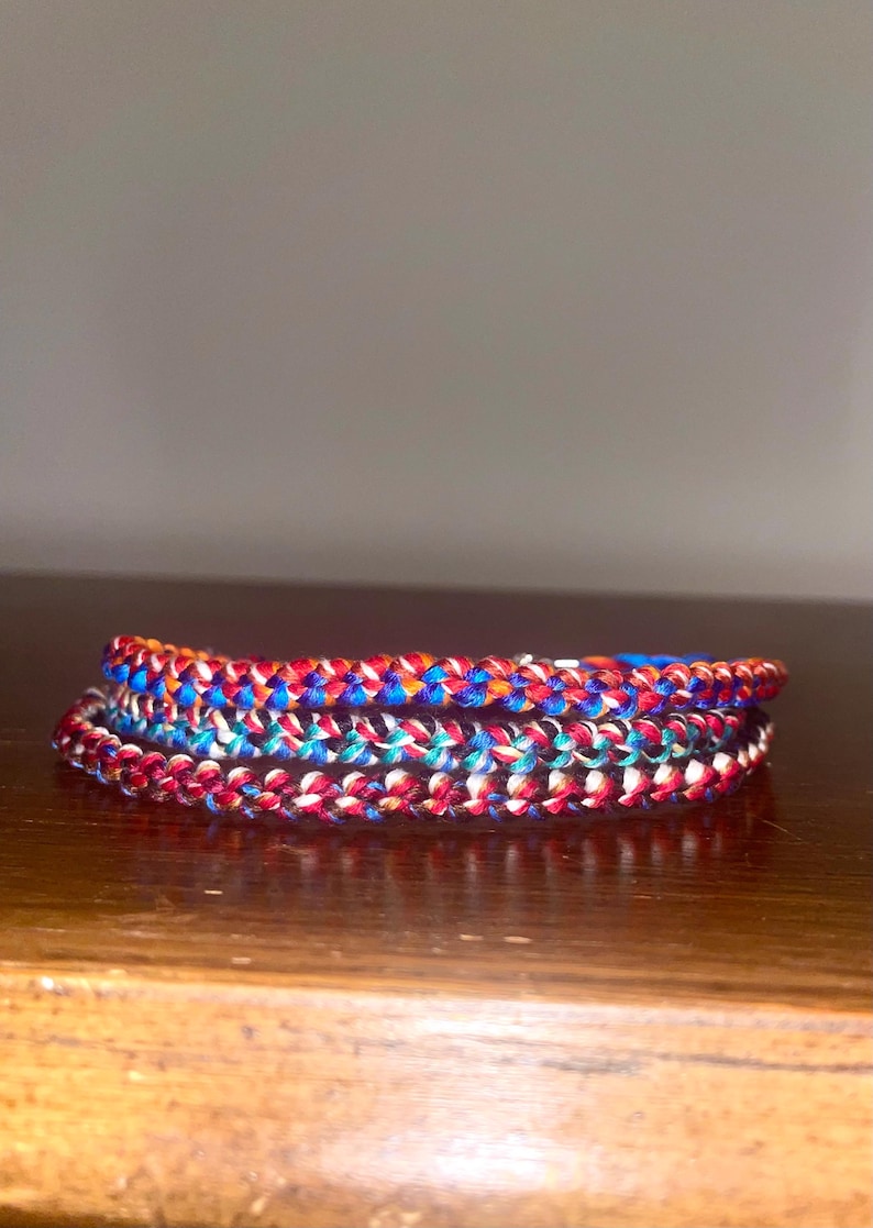 May include: A stack of colorful braided bracelets. The bracelets are made of red, white, blue, and orange threads. The bracelets are stacked on top of each other, creating a layered effect. The bracelets are on a wooden surface.