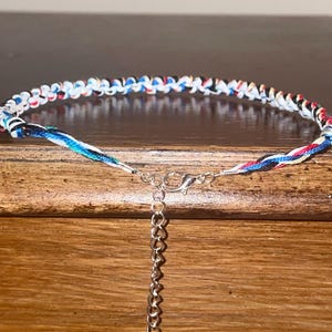 May include: A colorful braided bracelet with a silver-toned clasp and chain. The bracelet features a pattern of white, red, blue, and black threads. The bracelet is displayed on a wooden surface.