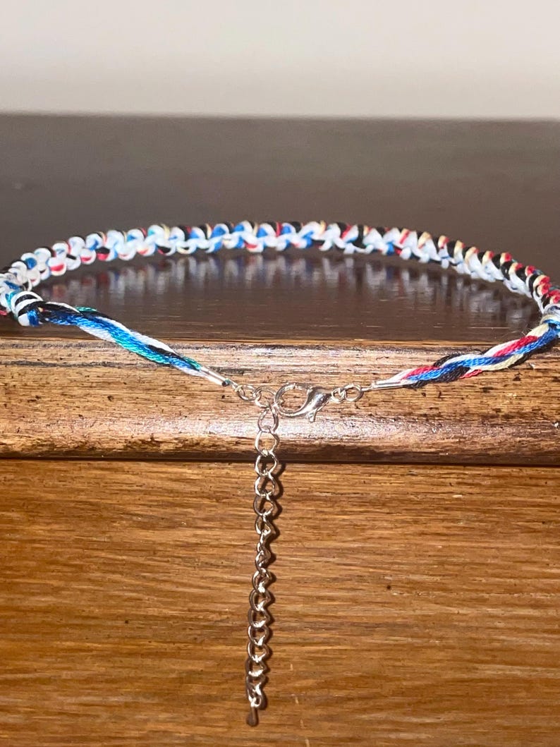 May include: A colorful braided bracelet with a silver-toned clasp and chain. The bracelet features a pattern of white, red, blue, and black threads. The bracelet is displayed on a wooden surface.